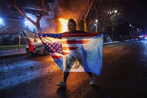 A person displaying multiple flags walks past a burning car during protests over the Trump administ…