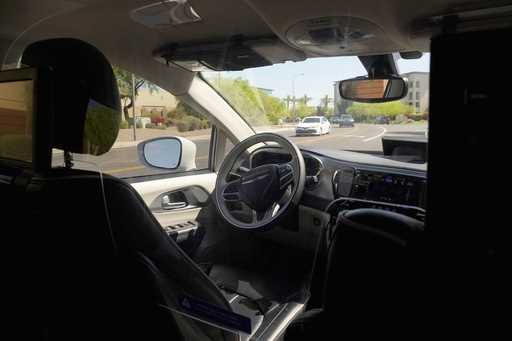 A Waymo minivan moves along a city street as an empty driver's seat and a moving steering wheel dri…