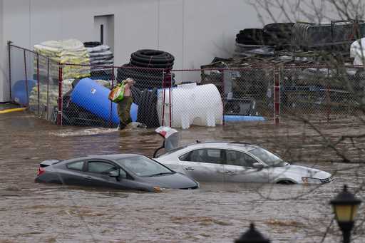 A man wearing chest waders walks past cars abandoned in floodwaters in a mall parking lot following…