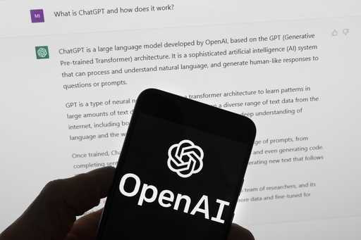 The OpenAI logo is seen on a mobile phone in front of a computer screen which displays output from …