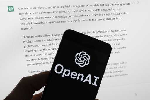 The OpenAI logo is seen on a mobile phone in front of a computer screen displaying output from Chat…