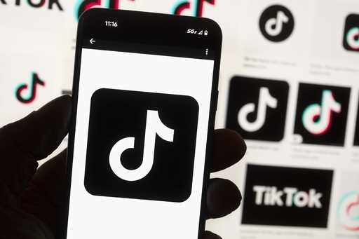 The TikTok logo is seen on a mobile phone in front of a computer screen which displays the TikTok h…