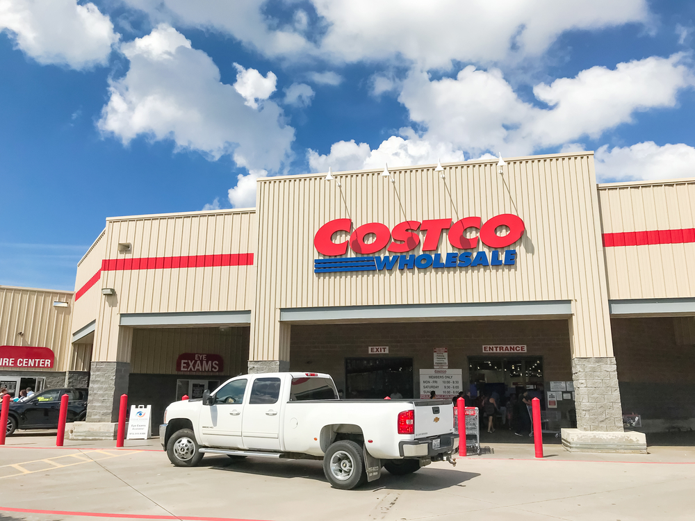 White truck driving by entrance facade of Costco Wholesale storefront. It is the largest membership-only warehouse club in USA, sunny summer cloud blue sky