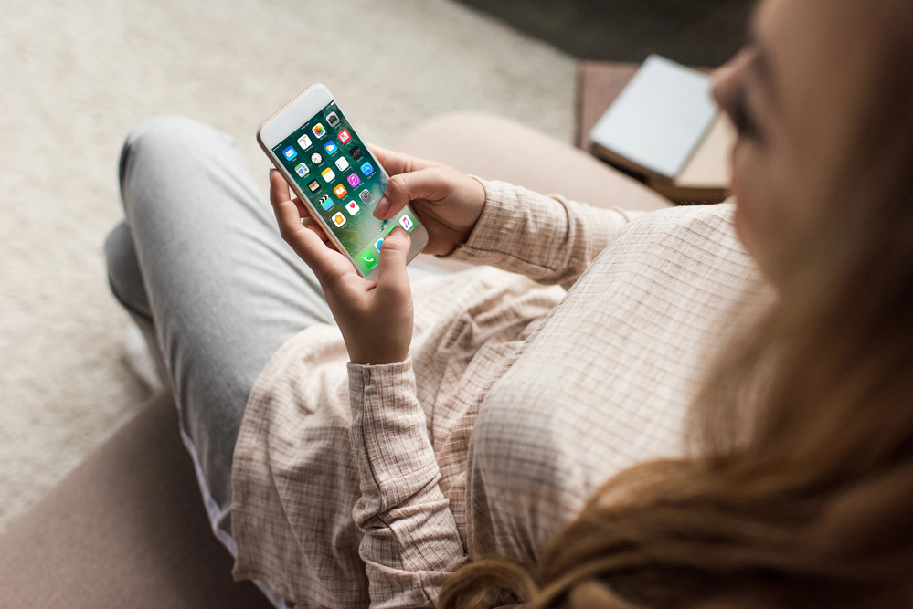 Cropped shot of woman on couch using smartphone with iOS apps on screen