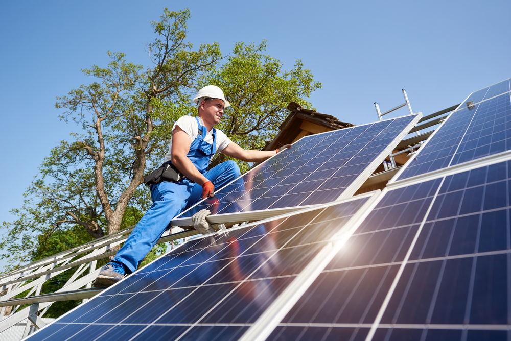 Young technician standing on metal platform installing heavy solar photo voltaic panel on blue sky background. Stand-alone solar panel system installation, efficiency and professionalism concept.