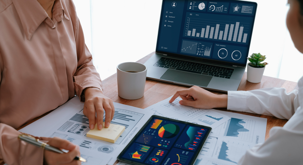 Analyst team colleague discuss financial data on digital dashboard, analyzing charts graph display on laptop and tablet screen. Modern office use business intelligence to plan marketing. Enthusiastic