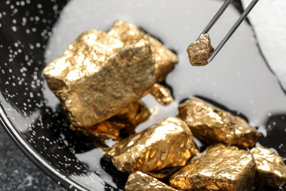 Tweezers with gold nugget above pile on grey table, closeup