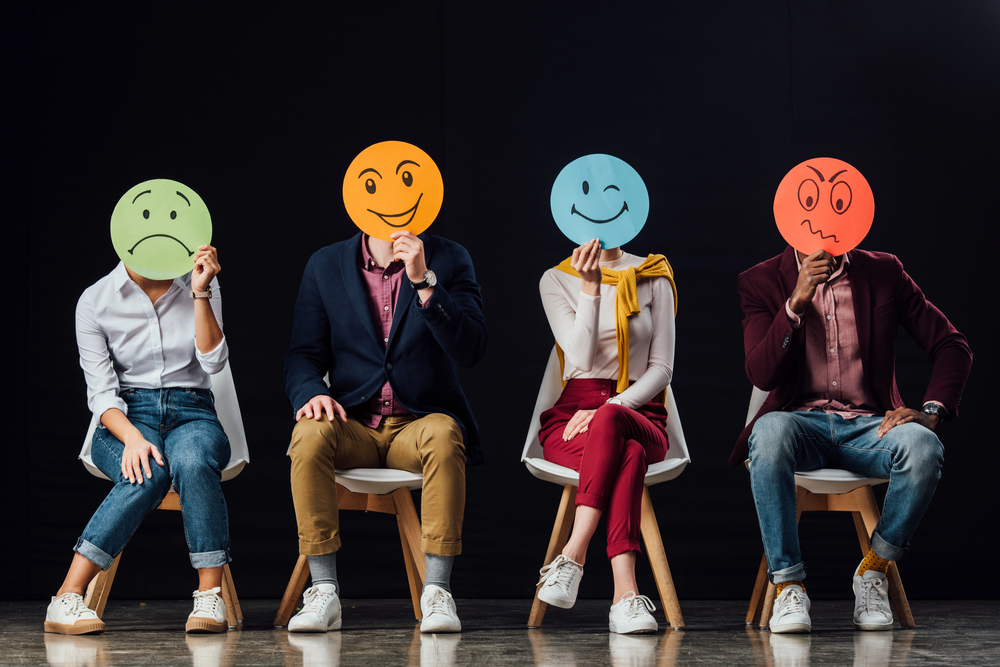 Photo of 4 people sitting on chairs holding up emotional faces drawn on construction paper.