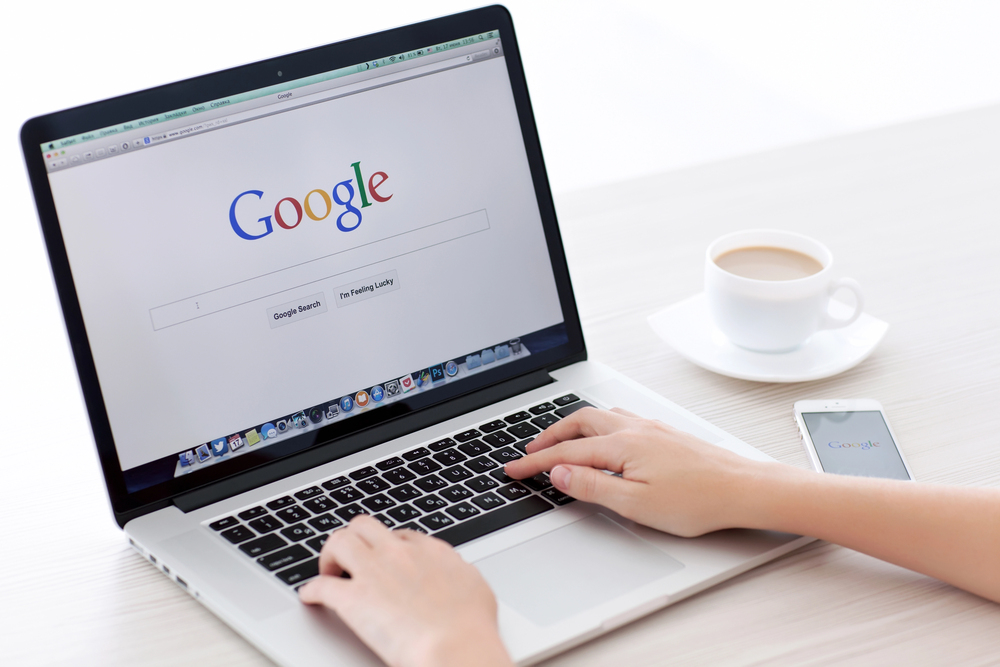 Photo of woman typing on a computer with Google logo on the screen and a coffee and phone next to her