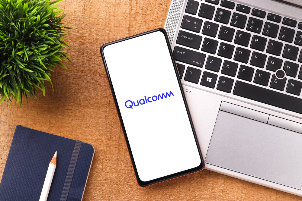 Photo of qualcomm logo on phone screen, phone sitting on a desk next to a laptop, notebook, pencil
