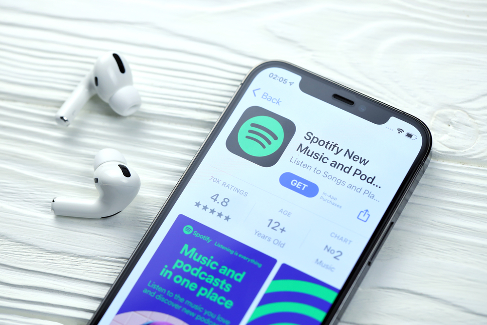 KHARKOV, UKRAINE - MARCH 5, 2021: Spotify icon and application from App store on iPhone 12 pro display screen with airpods pro on white wooden table - Stock Editorial Photography