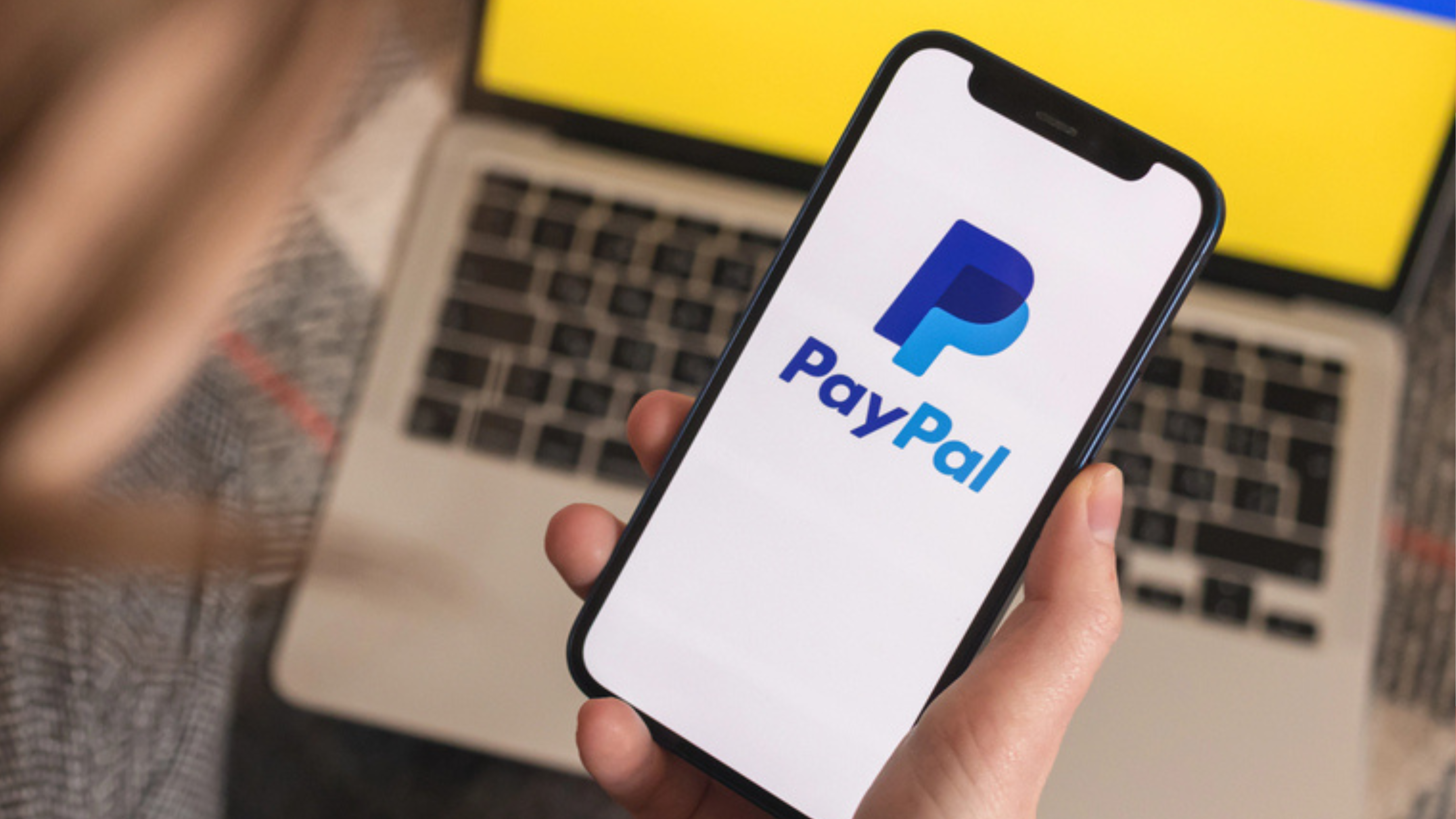 Kharkov, Ukraine - March 18, 2022: Young woman using Paypal in Ukraine. Mobile phone with logo and ukrainian flag on background