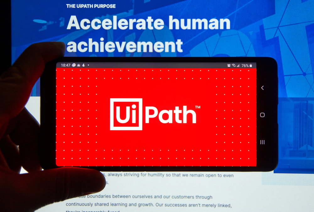 A hand holding a cellphone with UiPath logo over a laptop screen. UiPath is a global software company that develops a platform for robotic process automation.