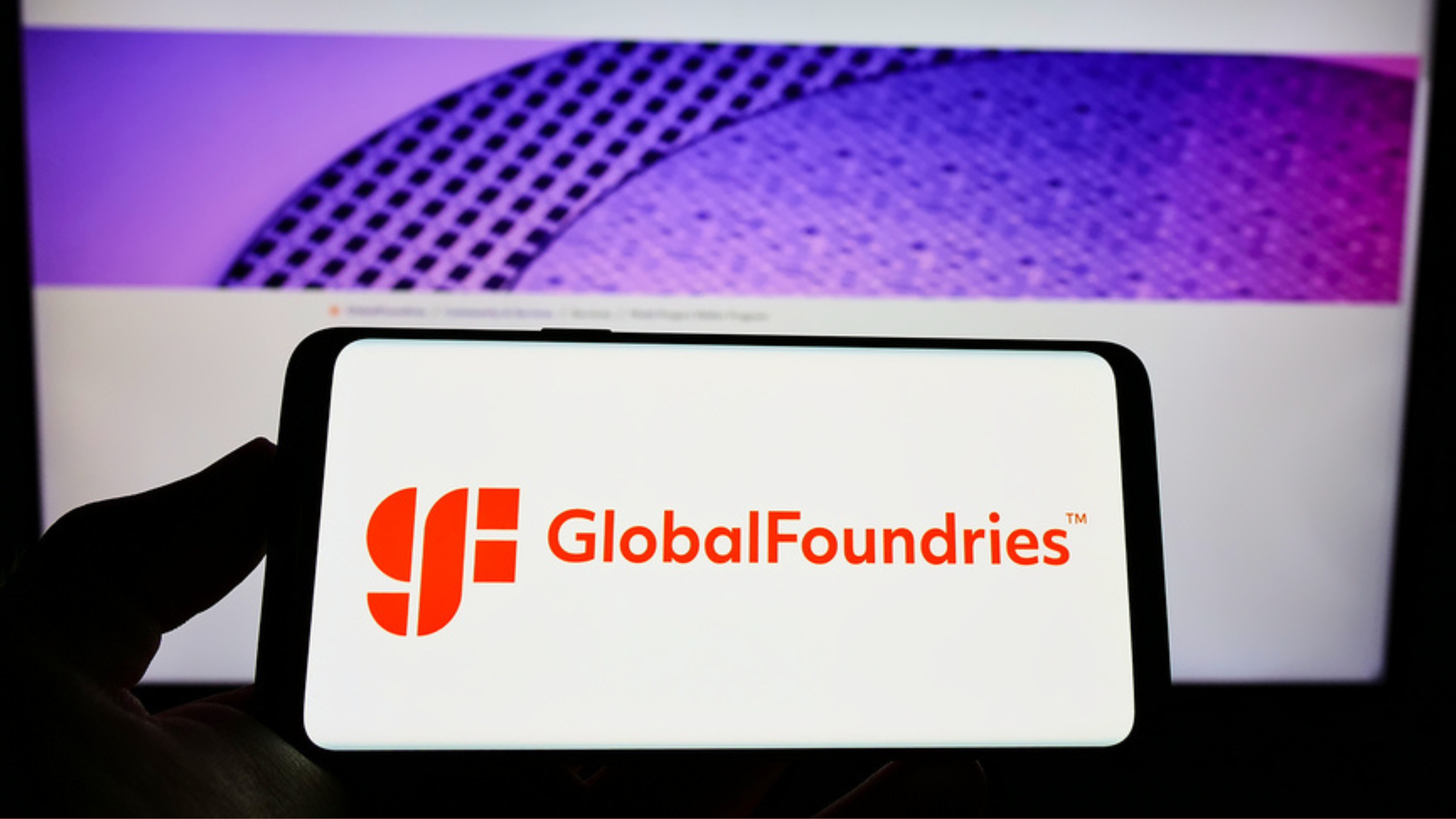 Person holding cellphone with logo of US semiconductor company GlobalFoundries Inc. (GF) on screen in front of webpage. Focus on phone display.