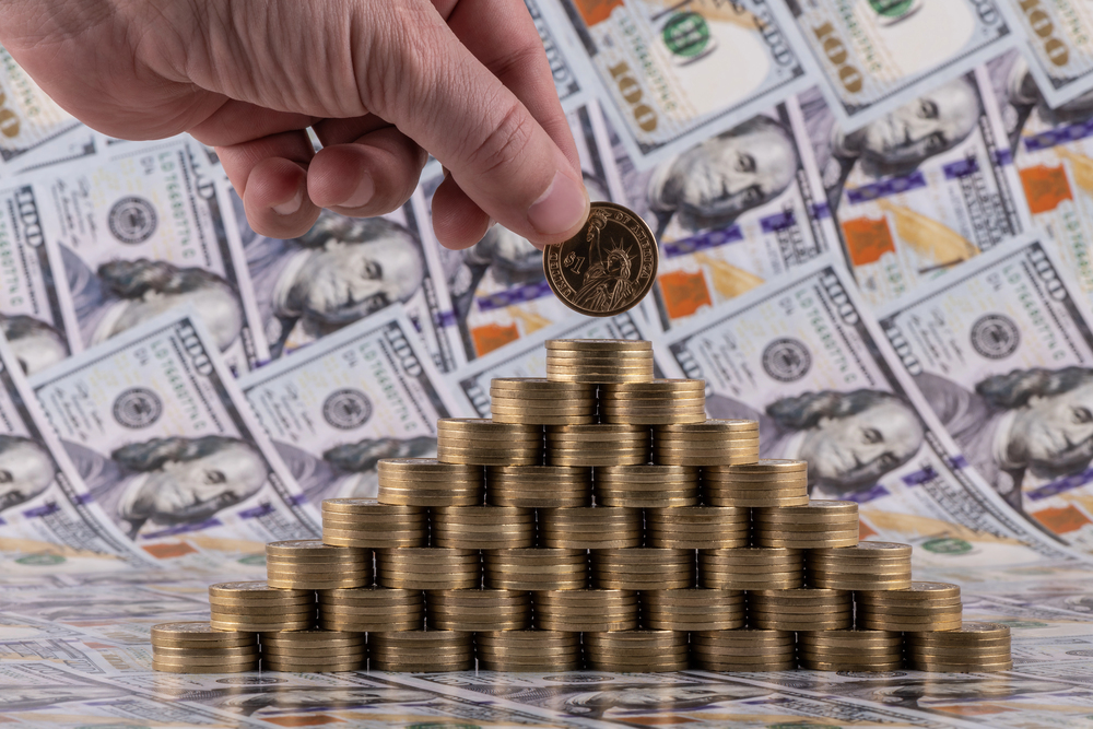 A man's hand puts a coin in denominations of 1 American dollar on top of a pyramid of gold coins against the background of bills in denominations of 100 US dollars — Photo
