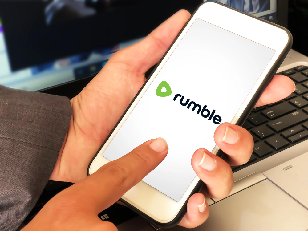 Man using video application Rumble on mobile phone with laptop in the backdrop.
