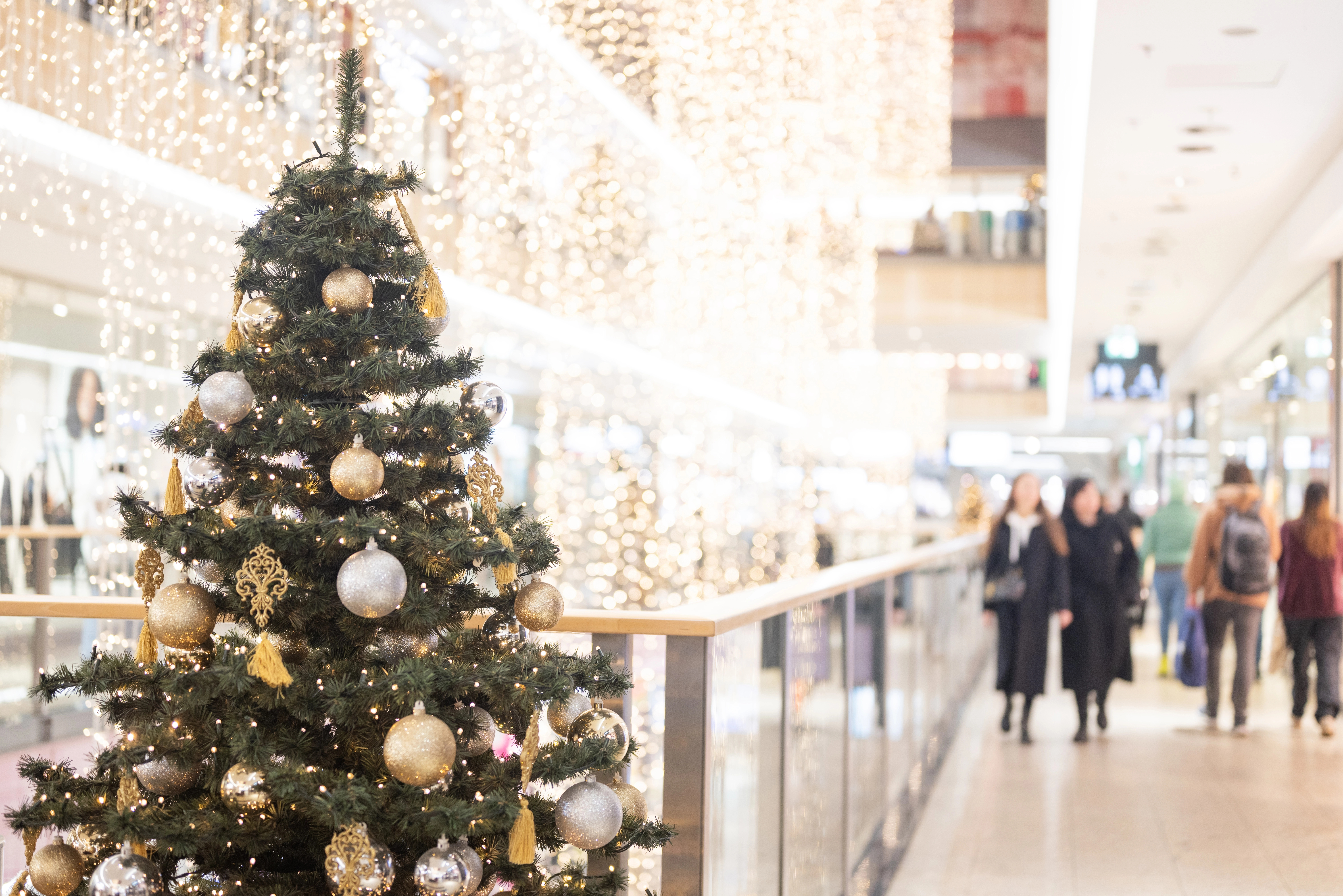 Decorated Christmas tree with gold and silver ornaments in a brightly lit shopping mall, with blurred shoppers in the background.