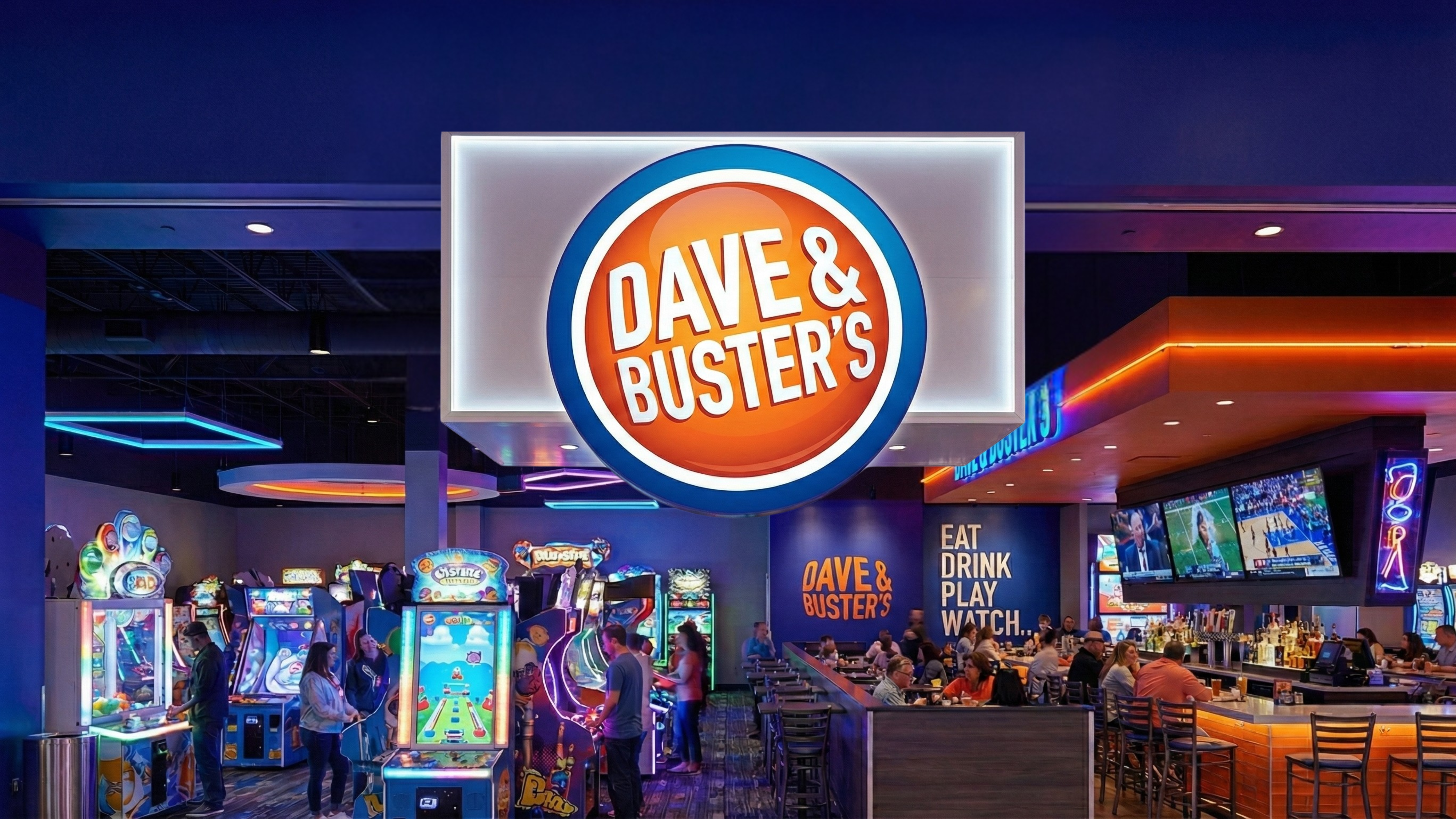Interior of a Dave & Buster’s with arcade games, dining area, and the large illuminated logo overhead.