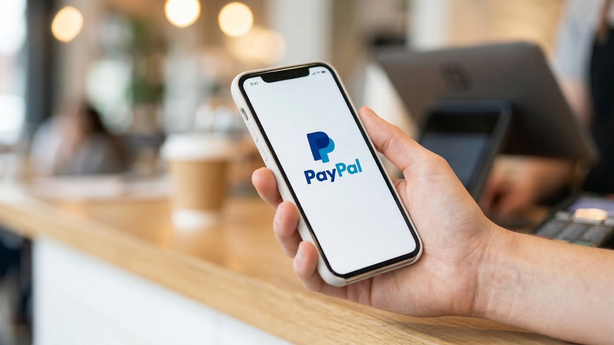 Customer holding smartphone displaying PayPal app at a checkout counter, highlighting digital payments platform.