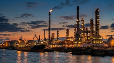 An illuminated oil refinery and docked tanker ship beside water at sunset.