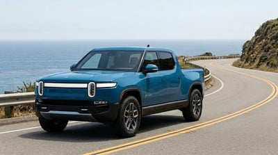 A blue Rivian R1T electric pickup truck drives along a coastal highway.