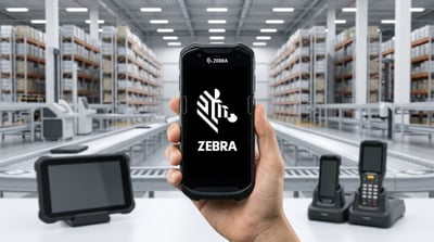 A hand holds a Zebra Technologies rugged mobile device displaying the Zebra logo inside a warehouse.