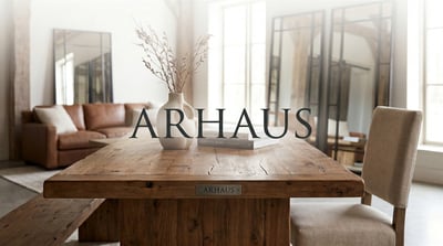 Arhaus logo overlaid on a branded rustic wood dining table in a furnished showroom setting.