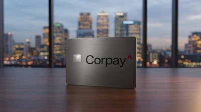 A metal Corpay-branded payment card stands upright on a wooden surface against a blurred city skyline backdrop.