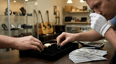 A pawnshop appraiser wearing white gloves examines jewelry on a velvet tray beside stacked US currency.