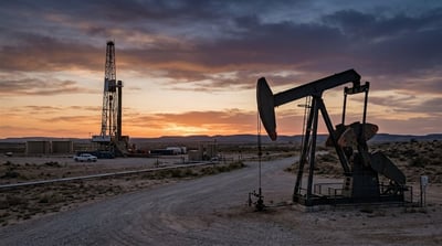 An oil pump jack and drilling rig stand on an arid plain at sunset.