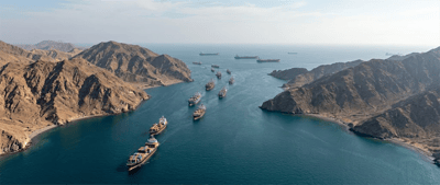 Multiple container ships and tankers navigate a narrow strait flanked by arid rocky mountains.