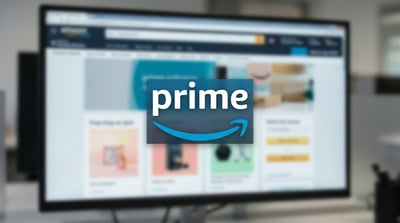 Amazon Prime logo overlaid on a blurred desktop monitor displaying the Amazon shopping website.