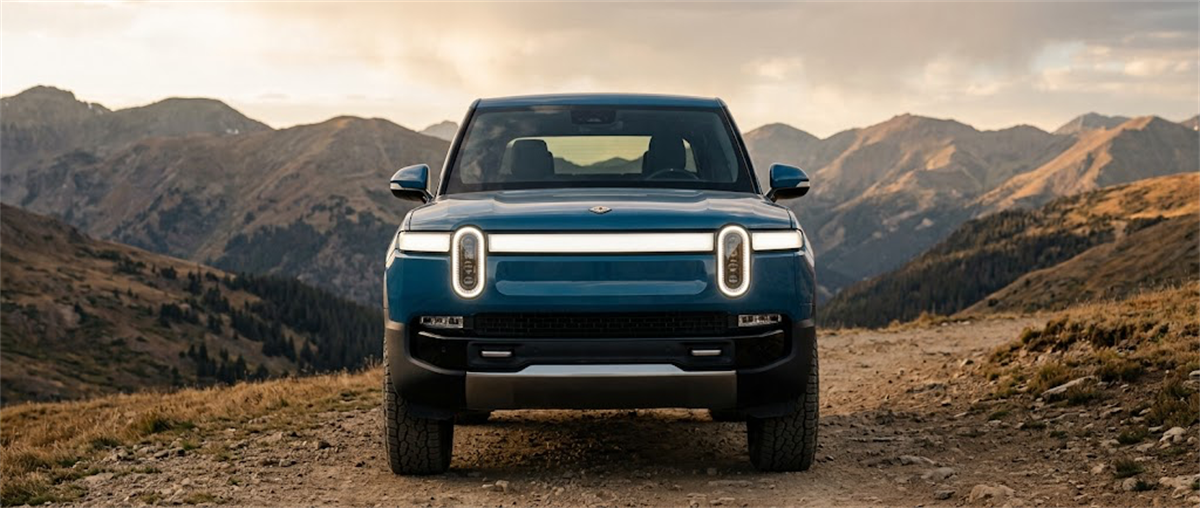 A blue Rivian electric truck faces forward on a dirt mountain road with rugged peaks in the background.