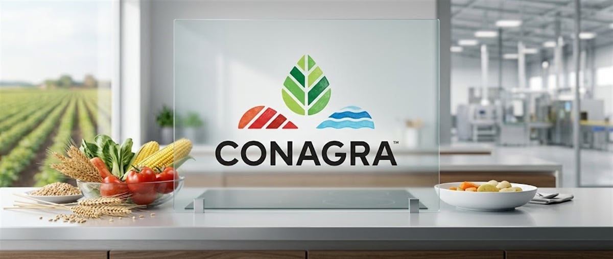 Conagra Brands logo on glass display surrounded by fresh vegetables, grains, and a blurred food processing facility.
