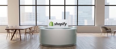 Shopify logo displayed on a sign at a round reception desk in a modern office with city views.
