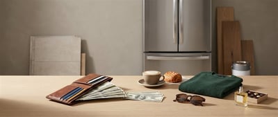 A wallet with cash, coffee, a sweater, sunglasses, and perfume on a counter beside a stainless steel refrigerator.
