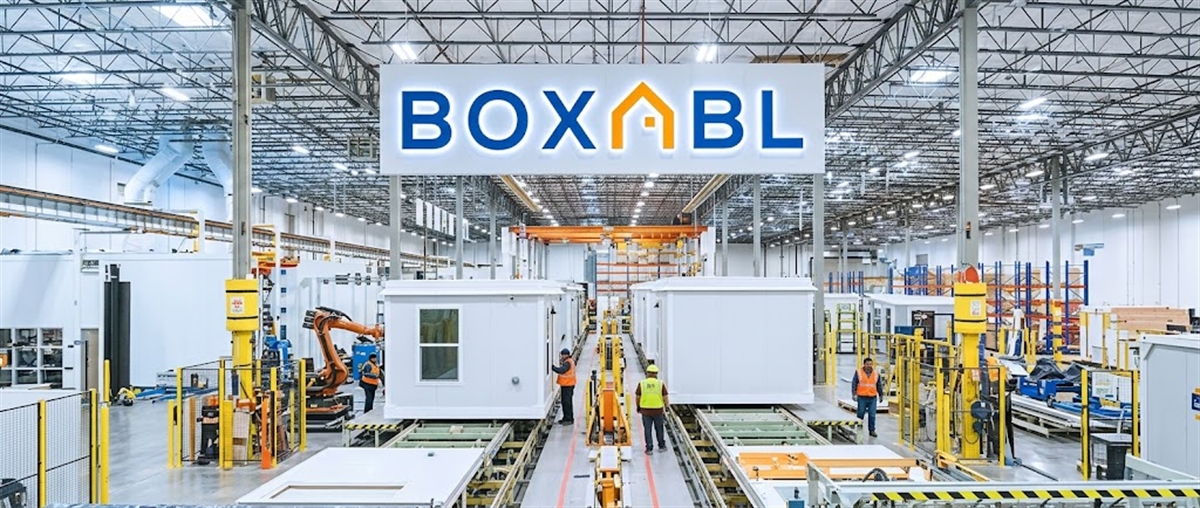 Boxabl logo hangs above a factory floor where workers assemble modular building units on a production line.