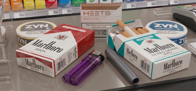 Marlboro cigarette packs, ZYN nicotine pouch tins, a HEETS tobacco box, a lighter, and a vape displayed on a store counter.
