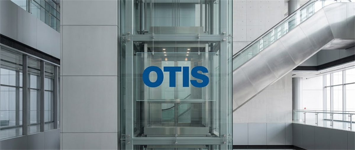 Glass elevator shaft displaying the Otis logo inside a modern building lobby with an escalator.