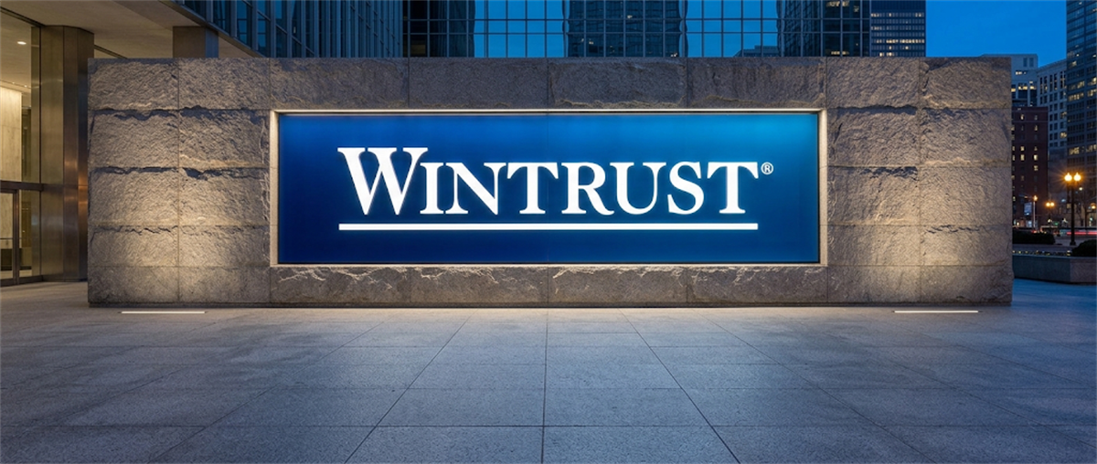 Illuminated Wintrust logo sign mounted on a stone wall outside an office building at dusk.