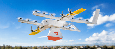 A DoorDash-branded delivery drone with a red and white cargo pod flies against a blue sky.