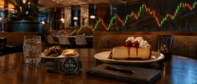 Restaurant table with cheesecake, dessert, water glass, smartwatch, and notebook overlaid with a stock candlestick chart.