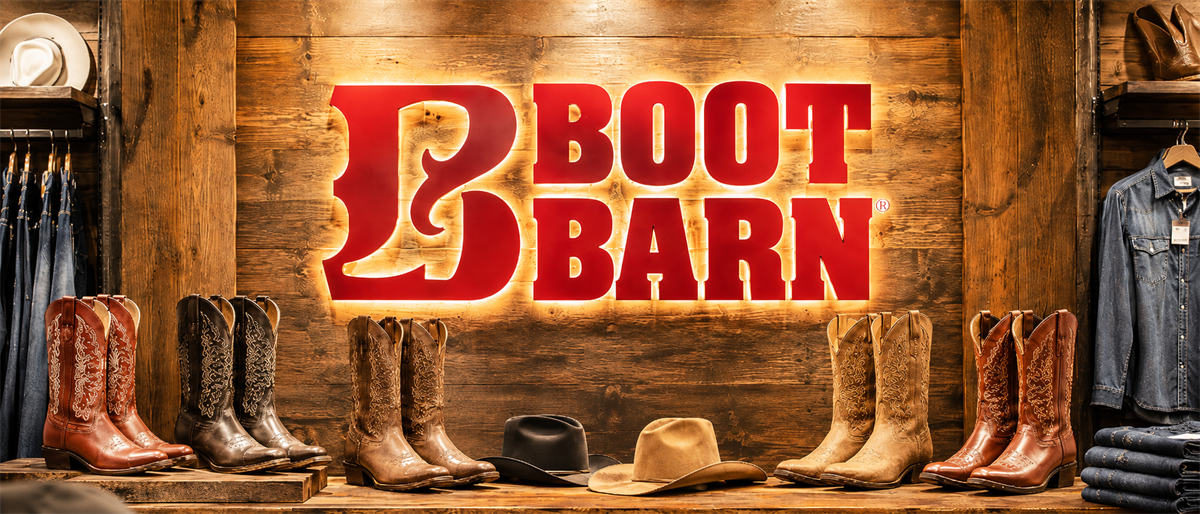 Boot Barn store display with illuminated logo, cowboy boots, hats, and denim clothing on wooden shelves.