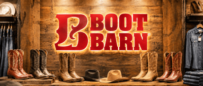 Boot Barn store display with illuminated logo, cowboy boots, hats, and denim clothing on wooden shelves.