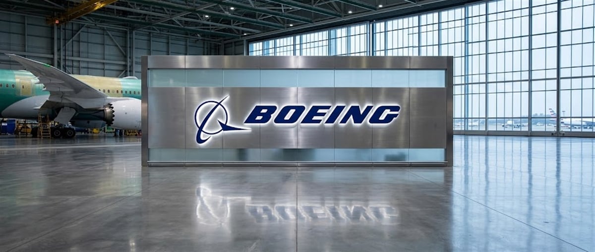 Boeing logo displayed on a metal wall inside an aircraft hangar with a partially assembled jet nearby.