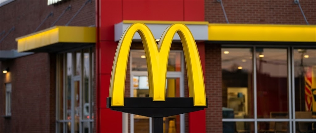 Golden Arches logo on a sign outside a McDonald's restaurant with brick facade and storefront windows.