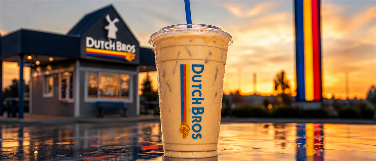 Dutch Bros iced coffee cup with logo in foreground of a Dutch Bros drive-thru location at sunset.
