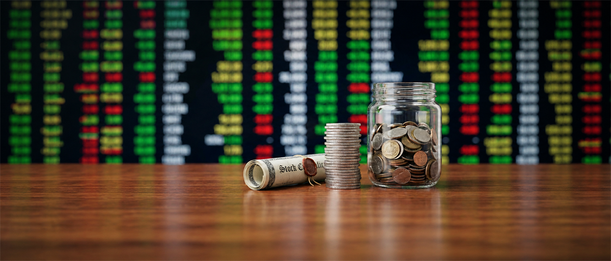 A rolled stock certificate, stacked coins, and a coin-filled jar sit on a table before a blurred stock ticker board.