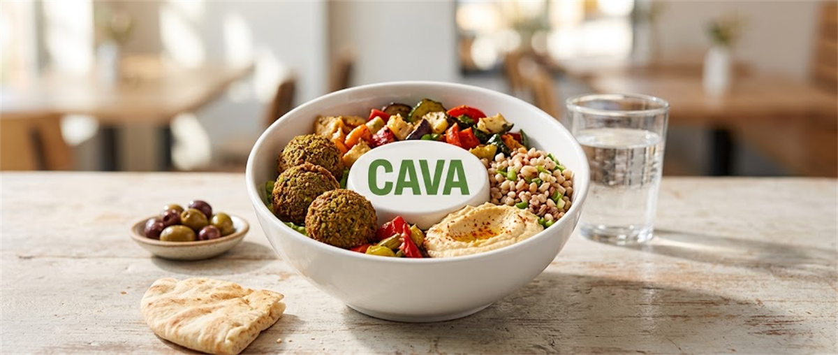 A CAVA-branded bowl of falafel, grains, and vegetables with hummus, olives, and pita on a restaurant table.