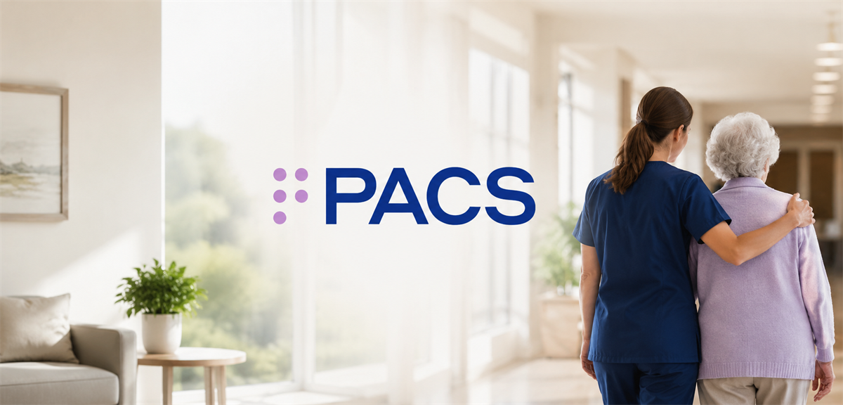PACS Group logo beside a caregiver walking arm in arm with an elderly woman in a care facility hallway.